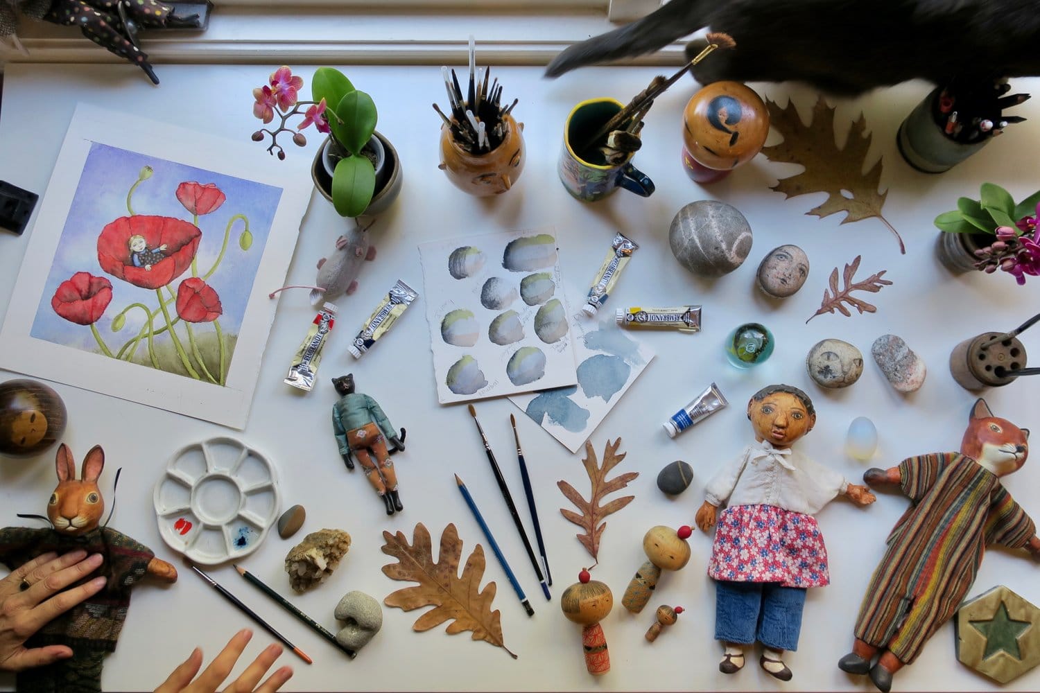 an artist's desk with paintings, painted rocks, puppets; you can see the artist's hands in the corner